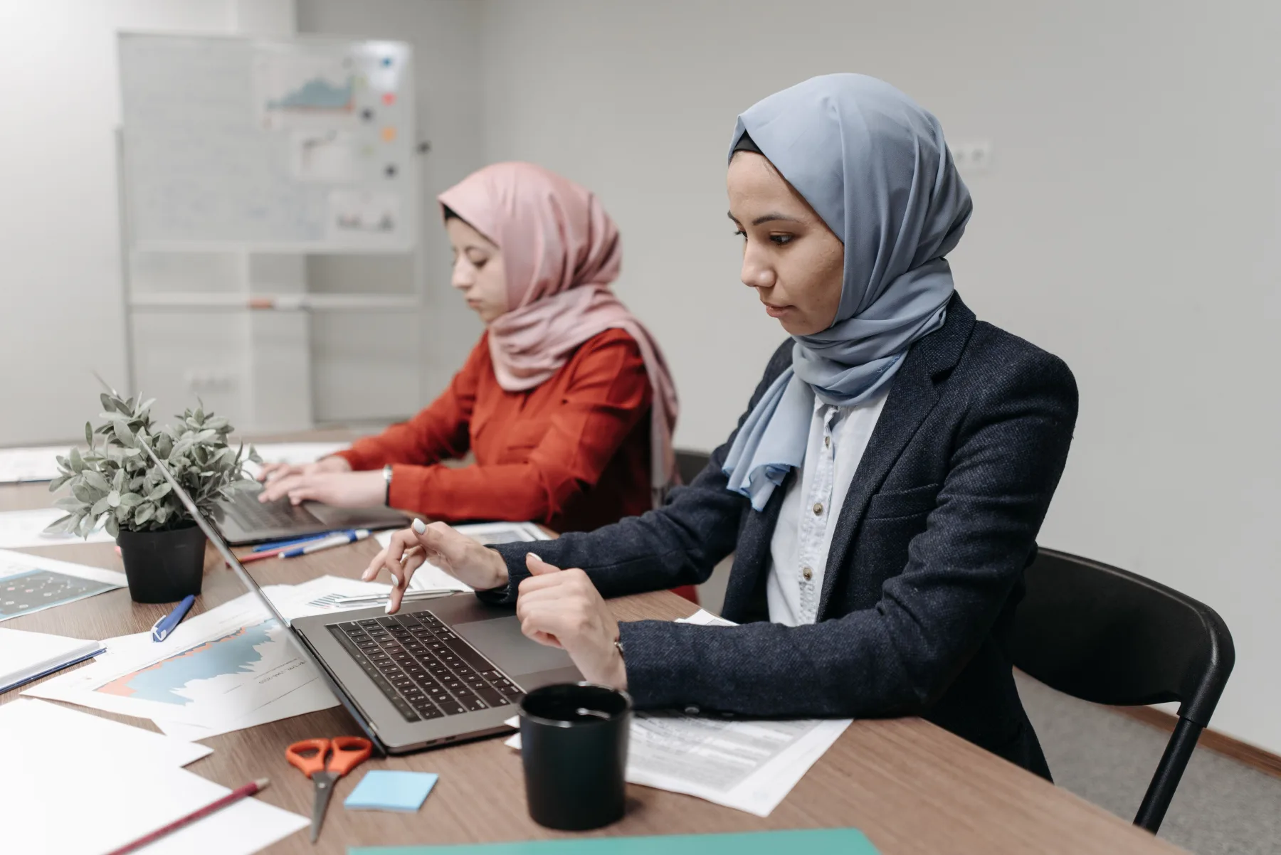 Dua perempuan berhijab bekerja dengan laptop dan laporan operasional